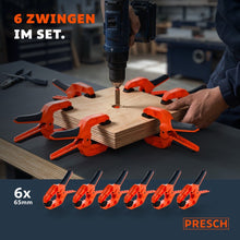 Sechs Federzwingen umschließen sternförmig ein Holzbrett, in dessen Mitte gerade gebohrt wird.